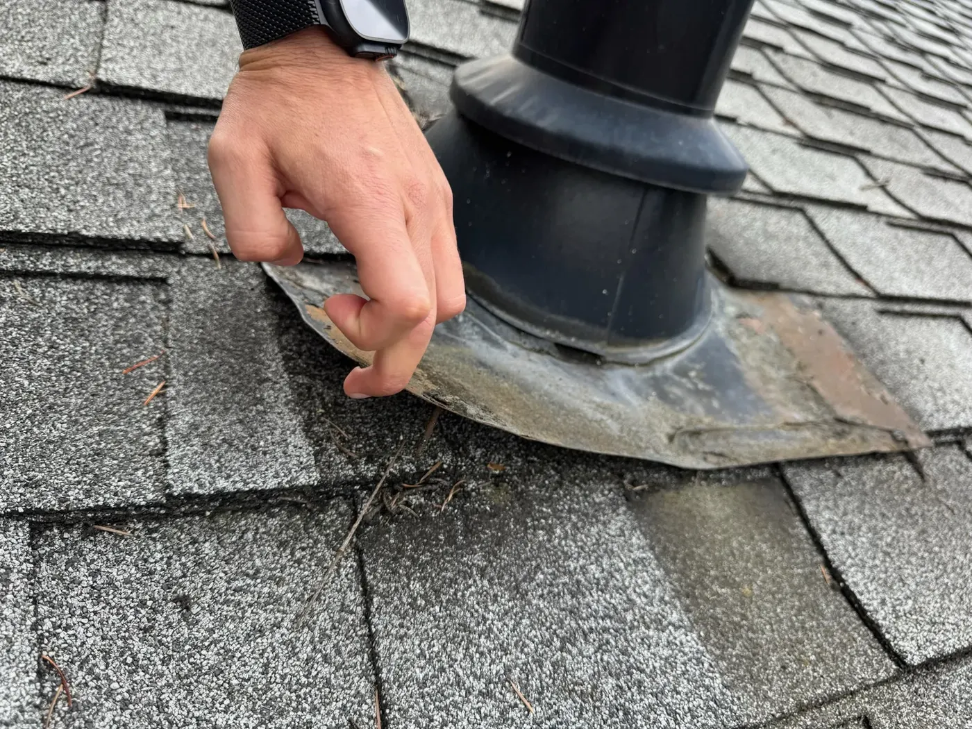 Roofer's hand inspecting a worn rubber pipe boot on a composition shingle roof in Seattle